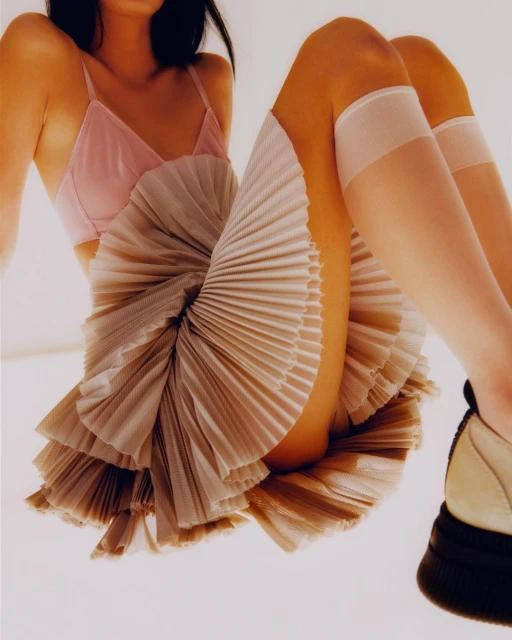 Woman in pink bra and tan pleated skirt sitting on white surface, showcasing fashion and style, with white knee-high socks and platform shoe.