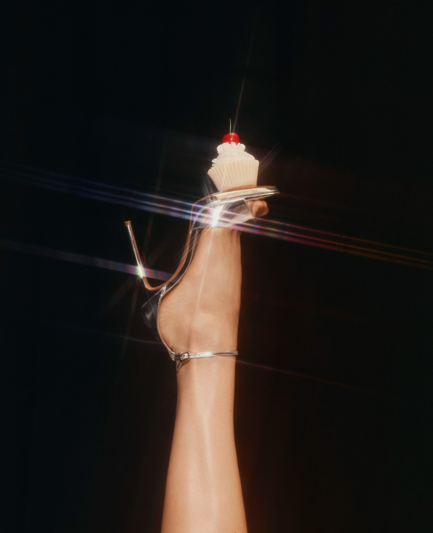 Elegant high-heeled shoe and a frosted cupcake with a cherry on top against a dark backdrop with visible light flares.