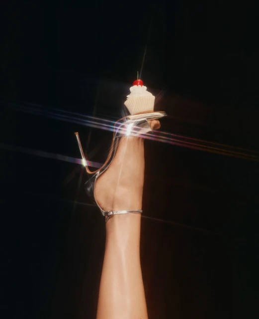 Elegant high-heeled shoe and a frosted cupcake with a cherry on top against a dark backdrop with visible light flares.