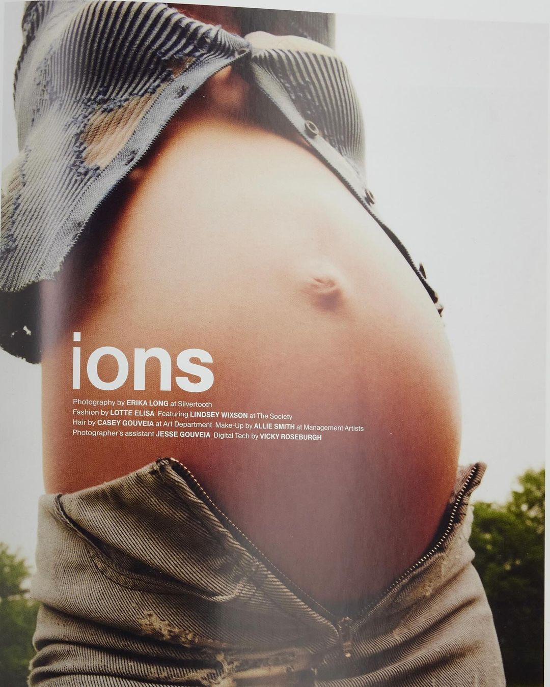Close-up studio shot of a pregnant adult female showing bare belly with open jeans and shirt against a blurred outdoor background.