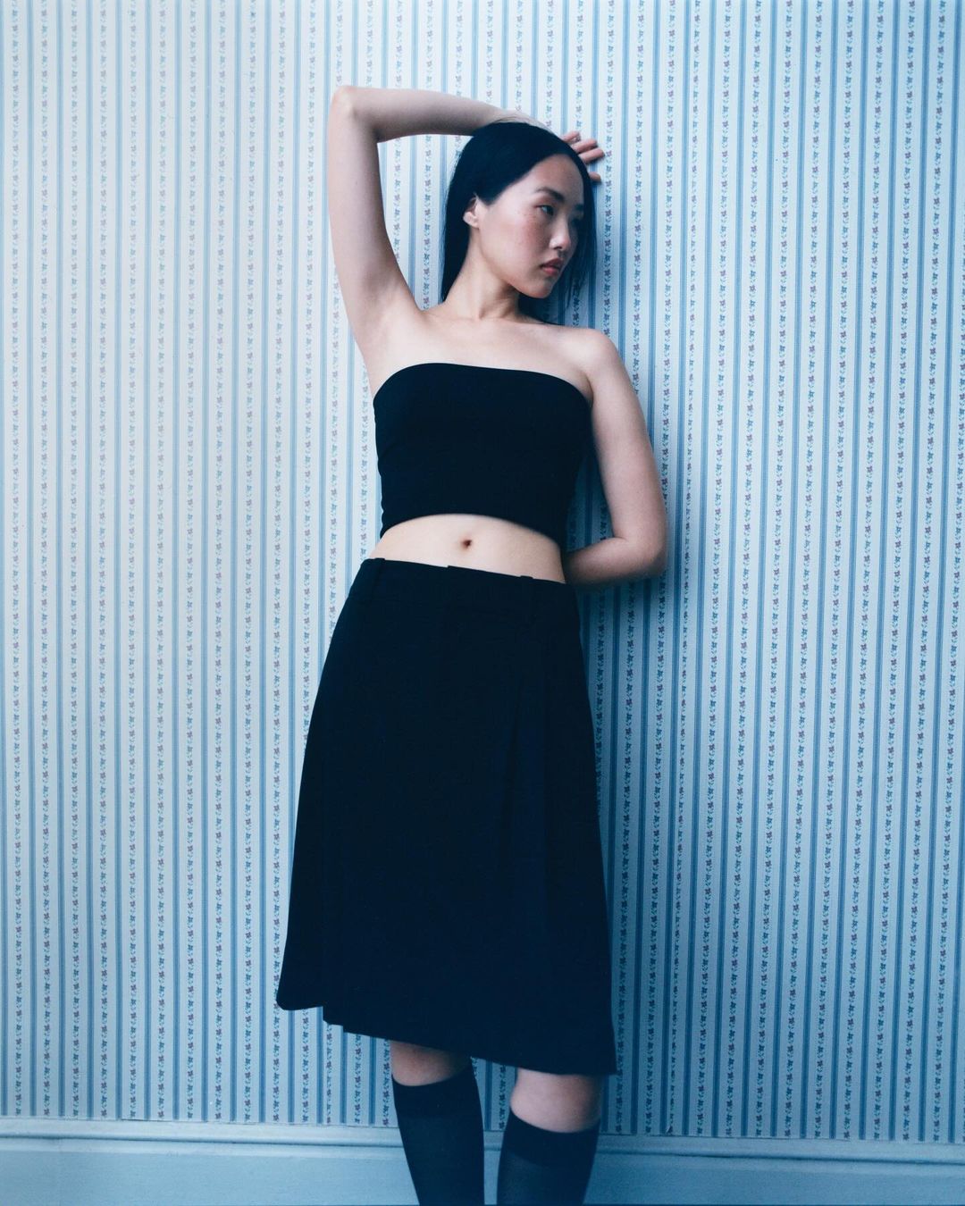 Young Asian woman poses in a black tube top and skirt against a blue and white striped wallpaper background in a fashion portrait.