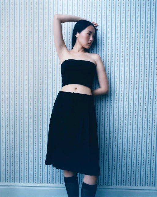 Young Asian woman poses in a black tube top and skirt against a blue and white striped wallpaper background in a fashion portrait.
