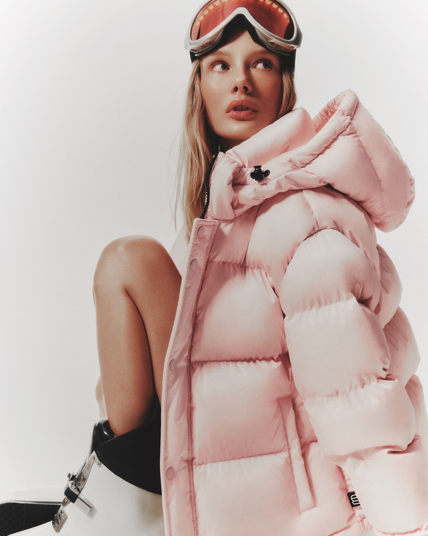 Young woman with ski goggles poses indoors wearing pink puffer jacket in a studio setting with a bright, neutral background.
