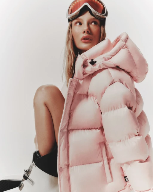 Young woman with ski goggles poses indoors wearing pink puffer jacket in a studio setting with a bright, neutral background.