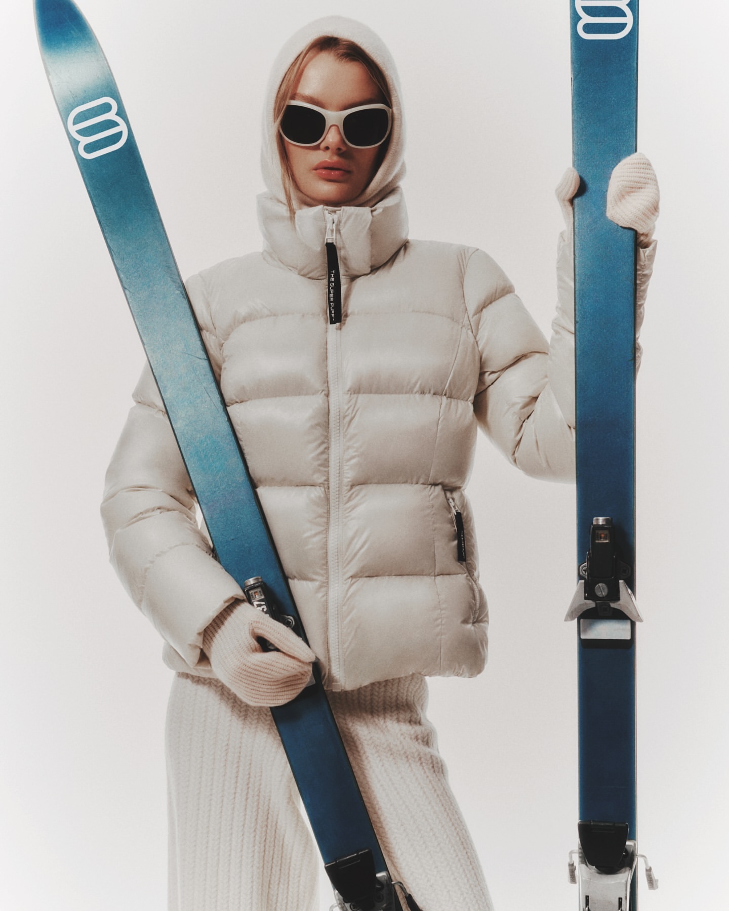 Blond woman poses in white puffer jacket, sunglasses, and knit gloves holding skis against a white backdrop for a winter fashion shoot.