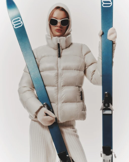 Blond woman poses in white puffer jacket, sunglasses, and knit gloves holding skis against a white backdrop for a winter fashion shoot.