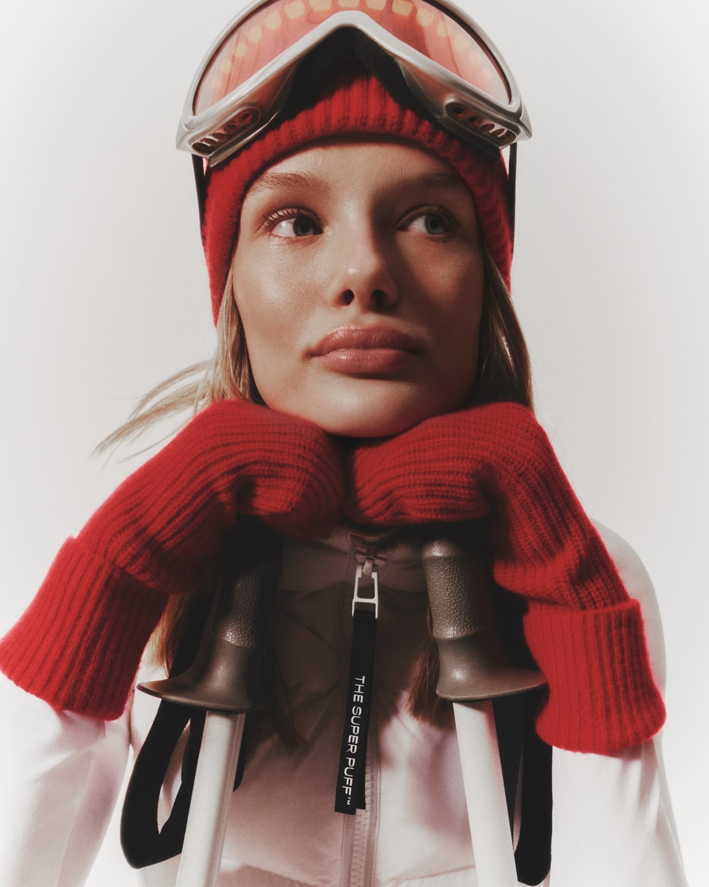 Close-up of a blonde adult female wearing a red beanie, ski goggles, and mittens, resting her chin on her hands, with ski poles.