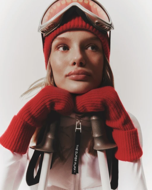 Close-up of a blonde adult female wearing a red beanie, ski goggles, and mittens, resting her chin on her hands, with ski poles.