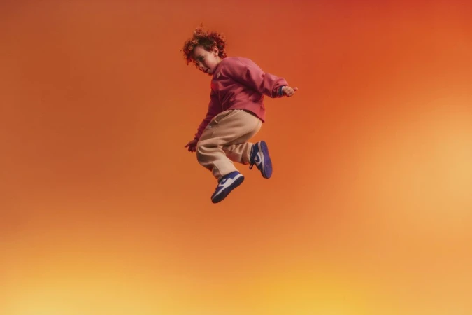 Young child with curly hair wearing a pink sweatshirt and beige sweatpants captured mid-air jumping against a warm orange gradient background.