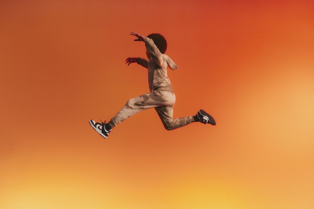 Young adult leaps mid-air, wearing a patterned hoodie and pants with black and white sneakers, set against an orange gradient backdrop.