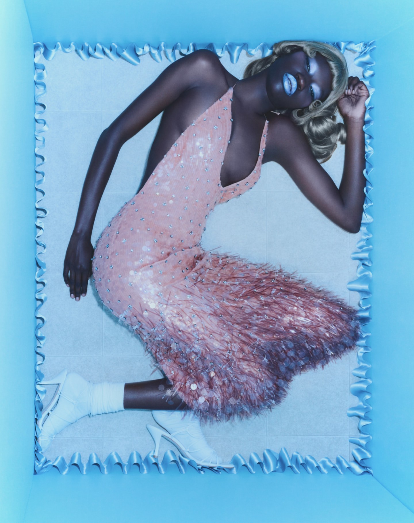 A woman with blond hair is reclined in a baby blue frame, wearing a pink embellished dress and white high heels in a studio setting.
