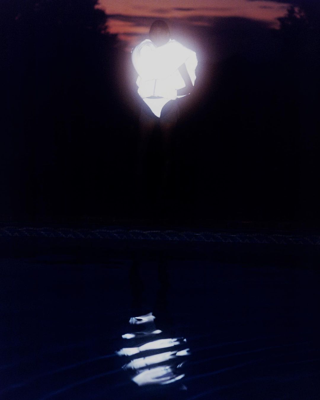 Figure in glowing white costume stands in dark landscape with water reflection. Dusk sky with orange and pink hues at horizon.