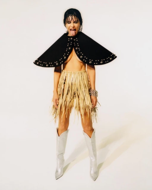 A woman with dark hair poses in a studio wearing a studded black cape, a raffia skirt, silver boots, and silver bracelets while sticking her tongue out.