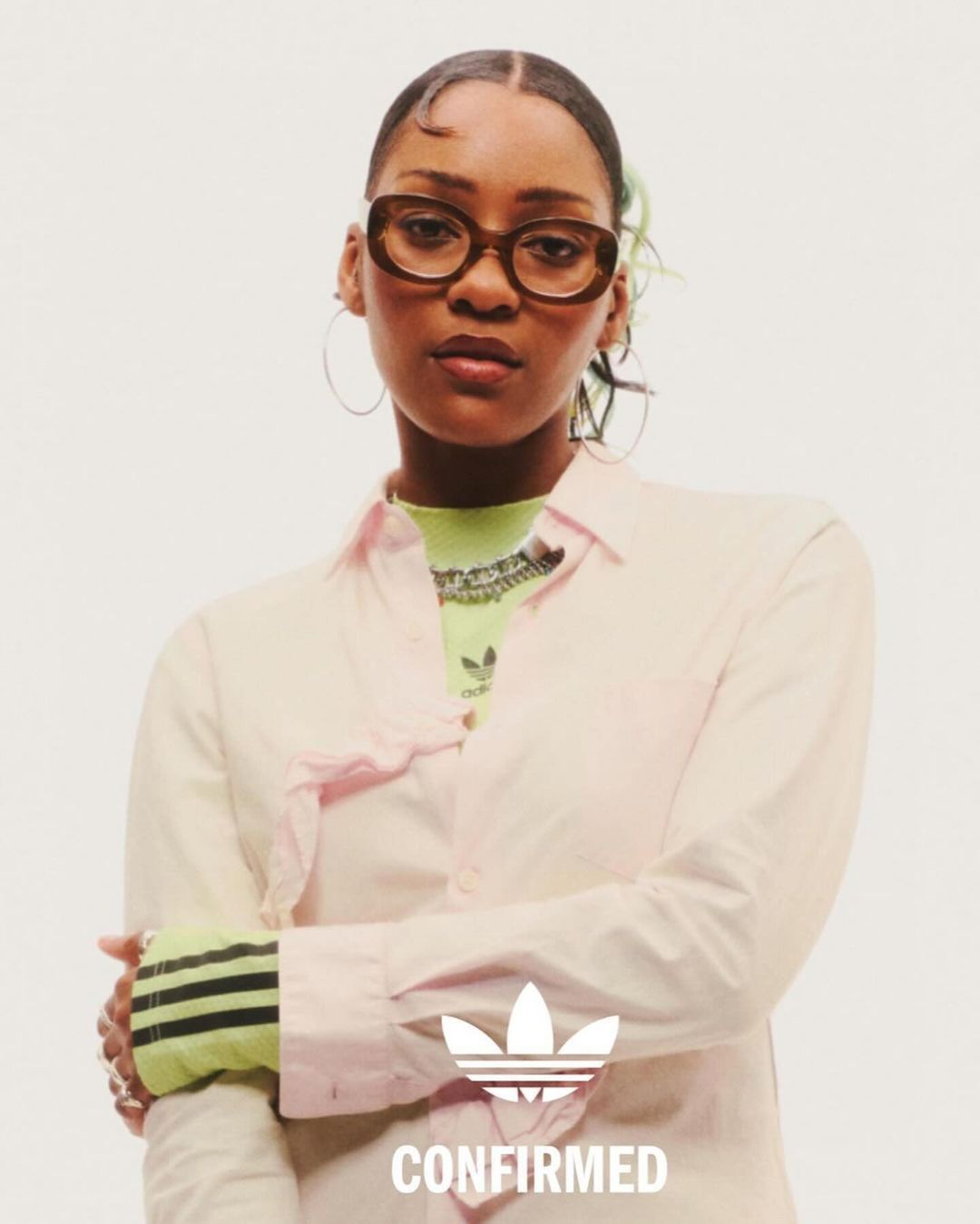 A woman poses wearing a light pink shirt and brown glasses, showcasing an Adidas logo. Close-up shot against a white background.