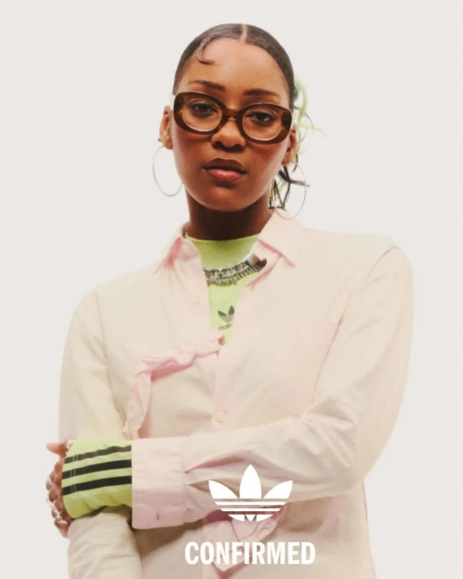 A woman poses wearing a light pink shirt and brown glasses, showcasing an Adidas logo. Close-up shot against a white background.