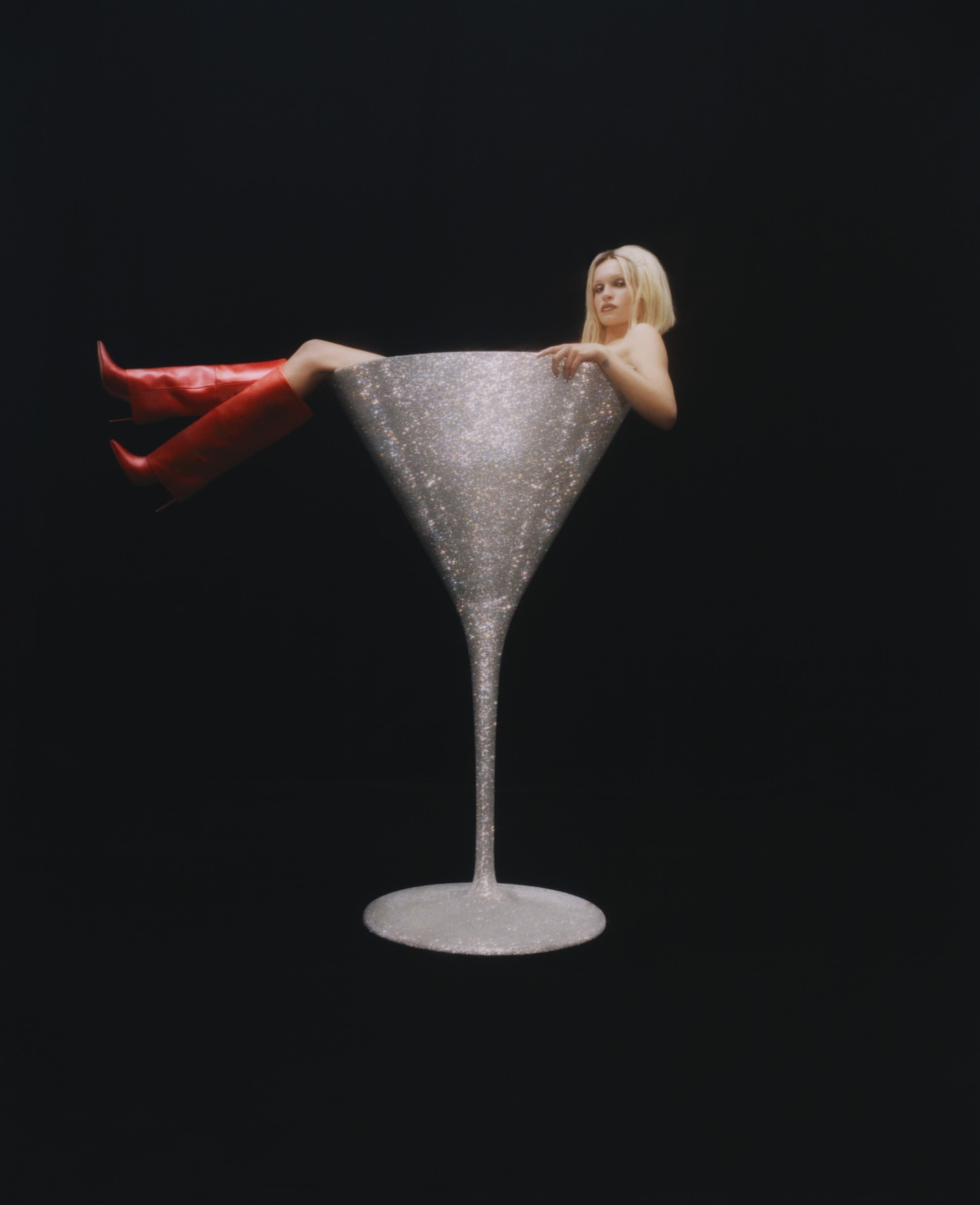 Blonde woman sitting inside a giant sparkly silver cocktail glass wearing red boots against a black background