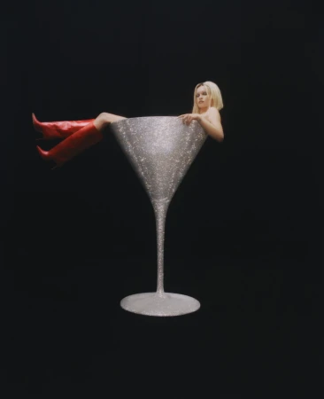 Blonde woman sitting inside a giant sparkly silver cocktail glass wearing red boots against a black background