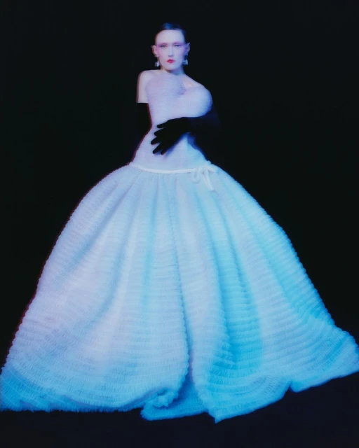 Fashion shot of a model wearing an elaborate light blue dress with a full skirt, with black opera gloves against a dark background.
