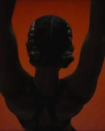 Rear view of a woman with braided hair wearing a black sports bra raising her arms against a dark orange background.