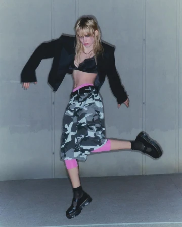 Blonde woman wearing a black blazer bralette and camouflage cargo shorts jumping indoors against a grey concrete wall