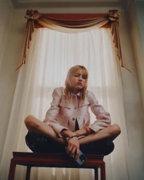 Low angle portrait of a young woman seated cross-legged on a dark wooden stool in front of a draped window by Erika Long