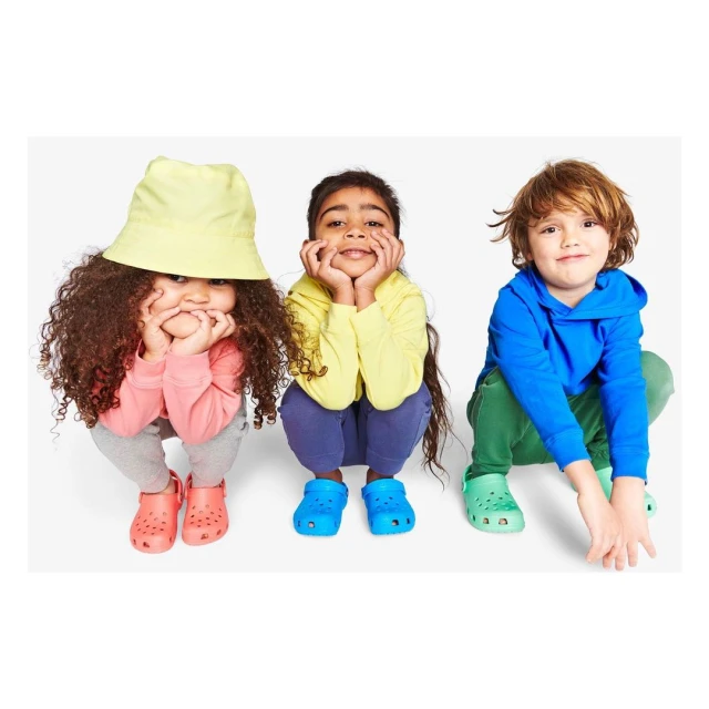 Three children squatting on a white surface, wearing colorful casual clothing and shoes, smiling, and looking directly at the camera.
