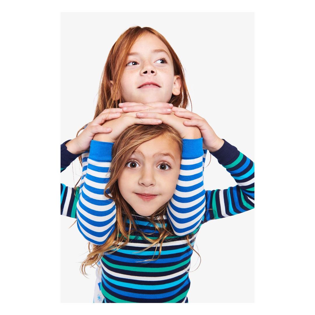Two smiling children pose in striped shirts against a white background. The redhead girl behind rests her hands on the blonde-haired girl.