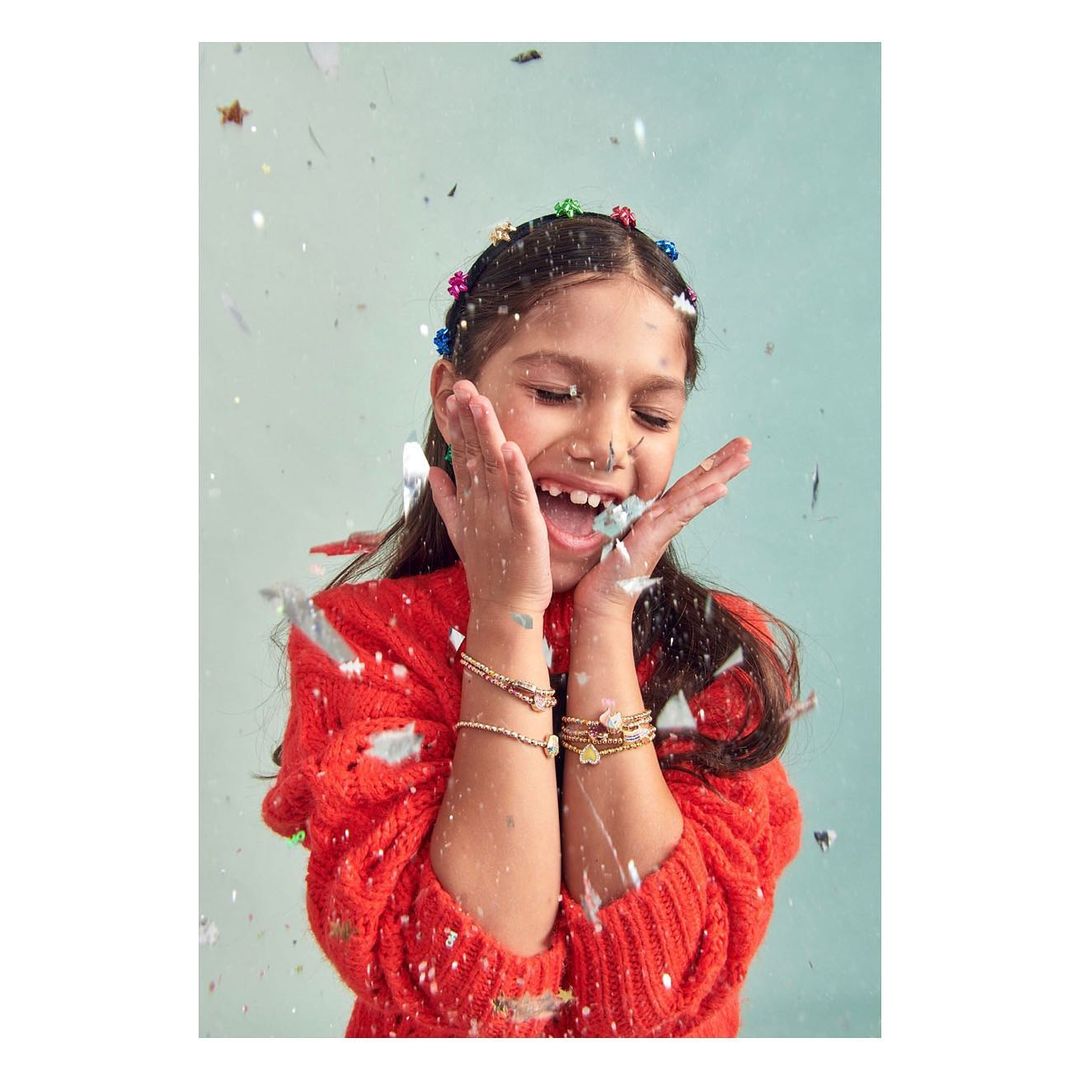 A cheerful young girl smiles with hands raised, surrounded by falling glitter against a light blue backdrop, wearing colorful hair clips.