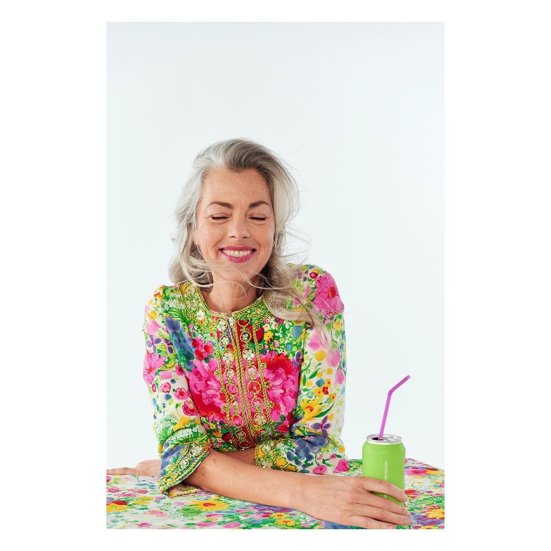 Smiling woman with gray hair in floral print jacket sits at floral table with green drink in can with pink straw against white backdrop.