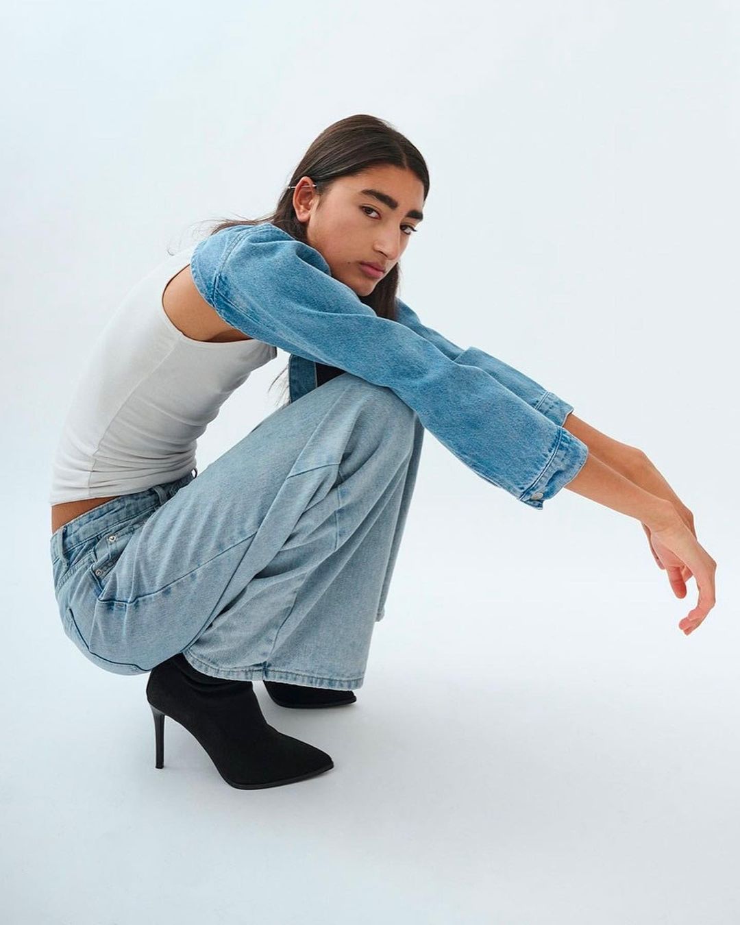 Woman in a light denim jacket and jeans squats in front of a white background, showcasing a casual yet stylish fashion look.