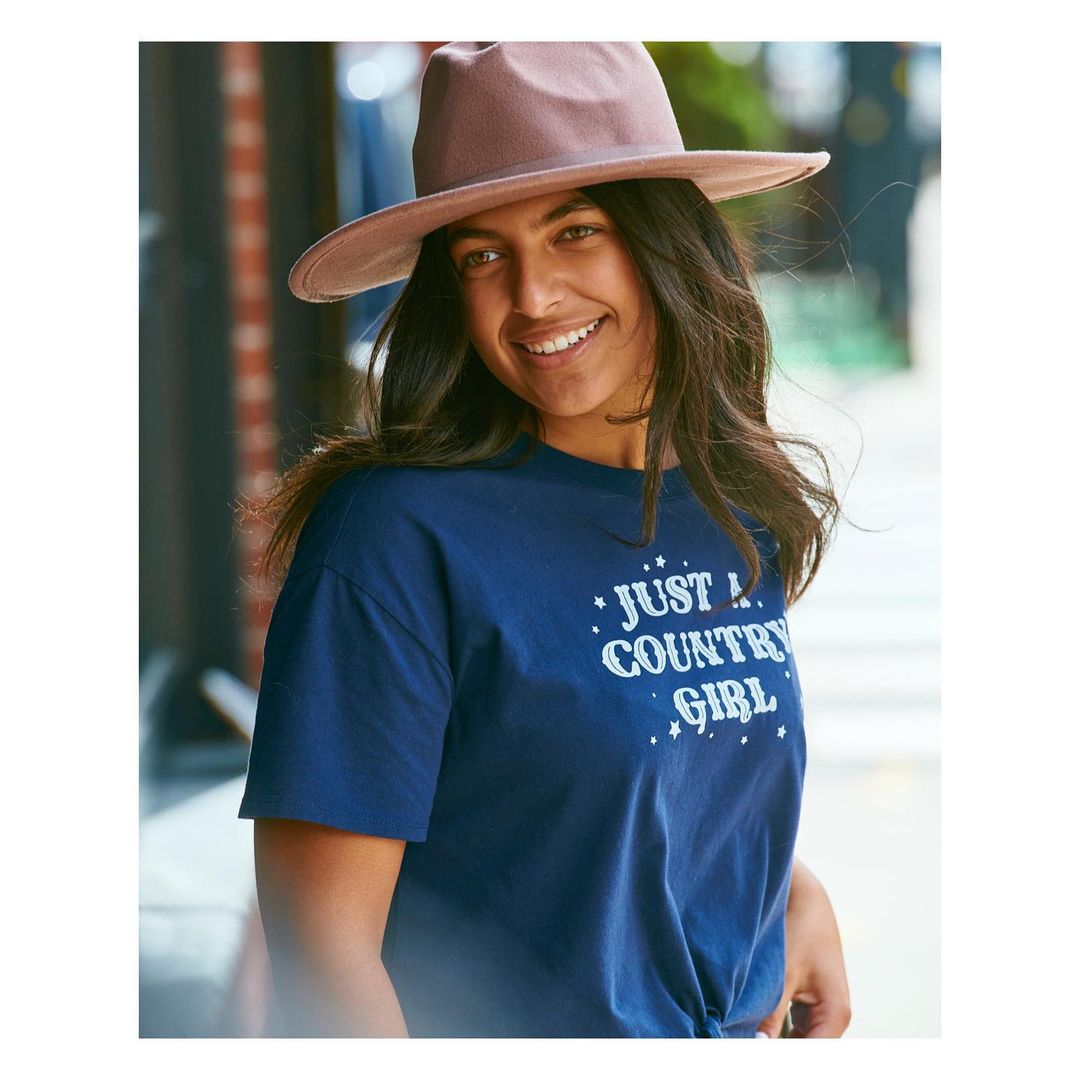 A smiling woman in a navy t-shirt and mauve hat stands outside with a brick wall and green foliage blurred in the background.
