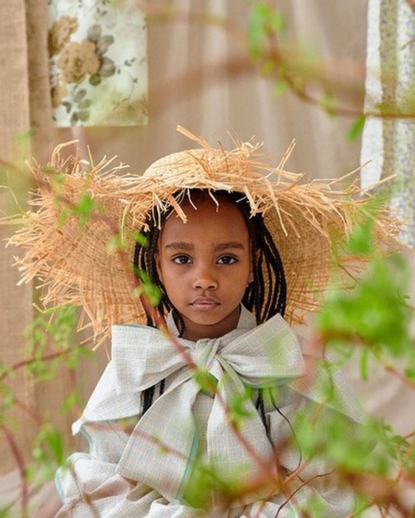Young Black child wearing a wide-brimmed straw hat and a light-colored bow-tied garment with out-of-focus foliage in the foreground.