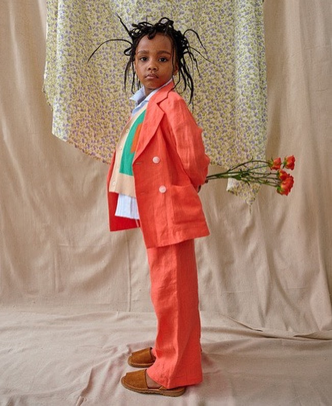 A young child dressed in a bright orange suit stands indoors holding roses against a neutral backdrop and floral-patterned fabric.