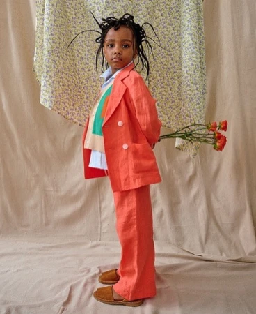 A young child dressed in a bright orange suit stands indoors holding roses against a neutral backdrop and floral-patterned fabric.