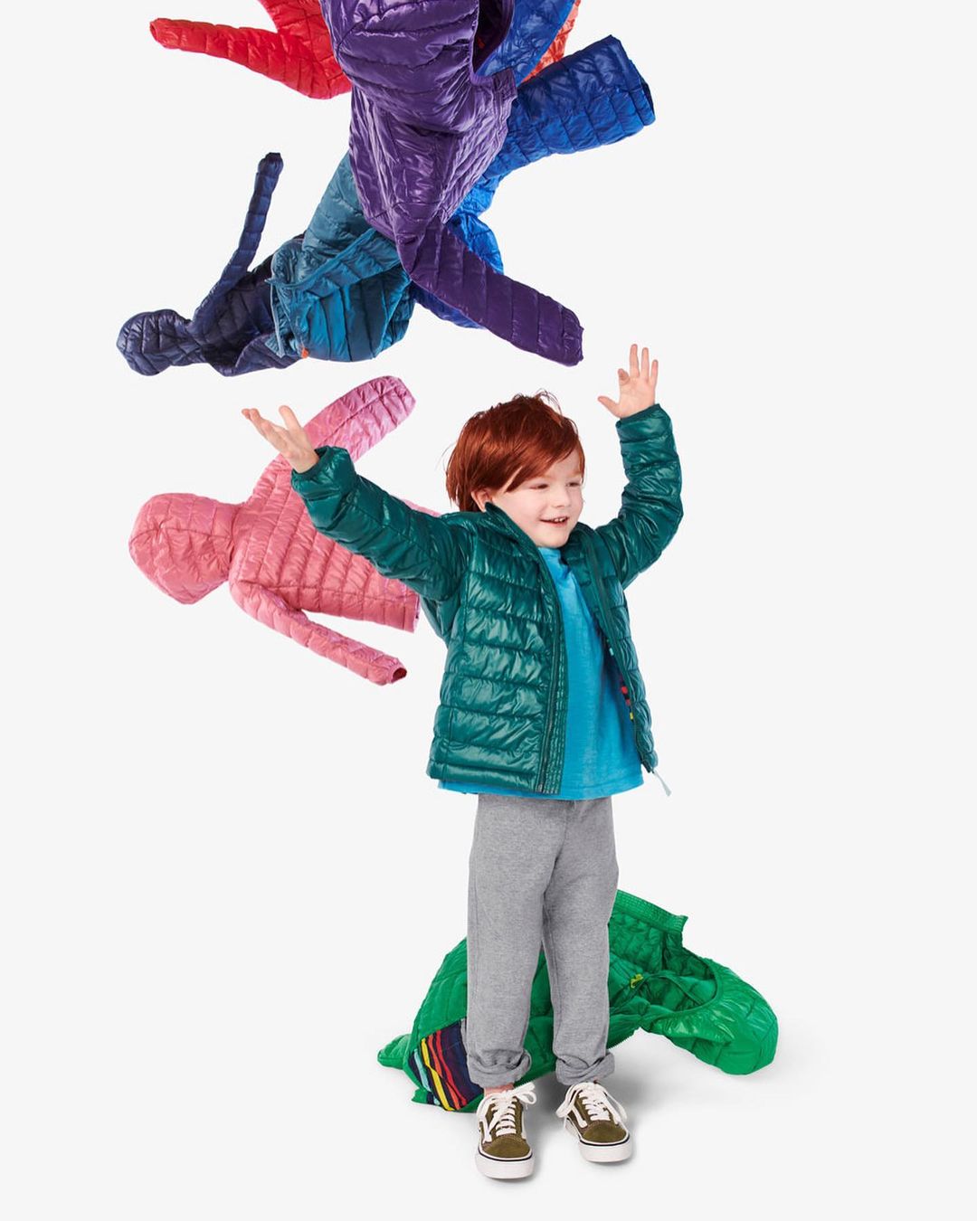 A young, red-haired child in a green jacket stands in a bright white studio with his arms raised as puffy coats float in the air.