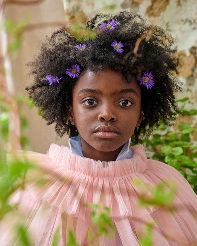 Portrait of a young black girl with afro hairstyle and flowers in her hair, wearing a frilly pink top, set against a floral backdrop.