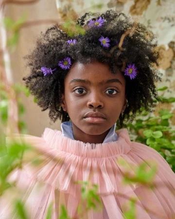 Portrait of a young black girl with afro hairstyle and flowers in her hair, wearing a frilly pink top, set against a floral backdrop.