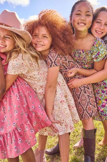 Group of five smiling young girls in patterned dresses, posing together outdoors in natural light, embracing and full of joy.