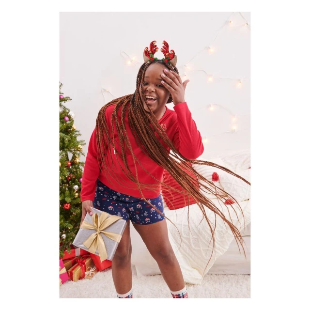 A smiling girl in a red holiday-themed shirt and festive shorts holds a gift in an indoor setting with a Christmas tree.