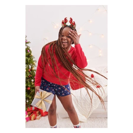 A smiling girl in a red holiday-themed shirt and festive shorts holds a gift in an indoor setting with a Christmas tree.