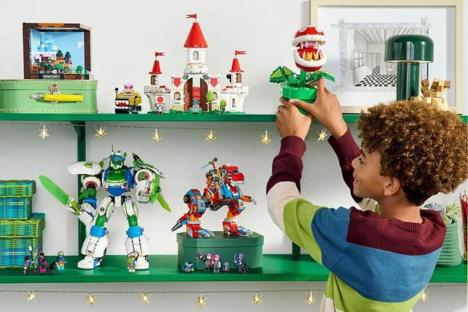 A boy places a Piranha Plant Lego set on a green shelf among other assembled sets in a brightly lit indoor setting.