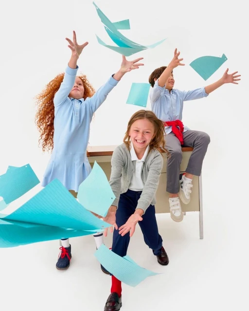 Three smiling children toss blue paper sheets in the air in a bright studio, creating a joyful and energetic scene.