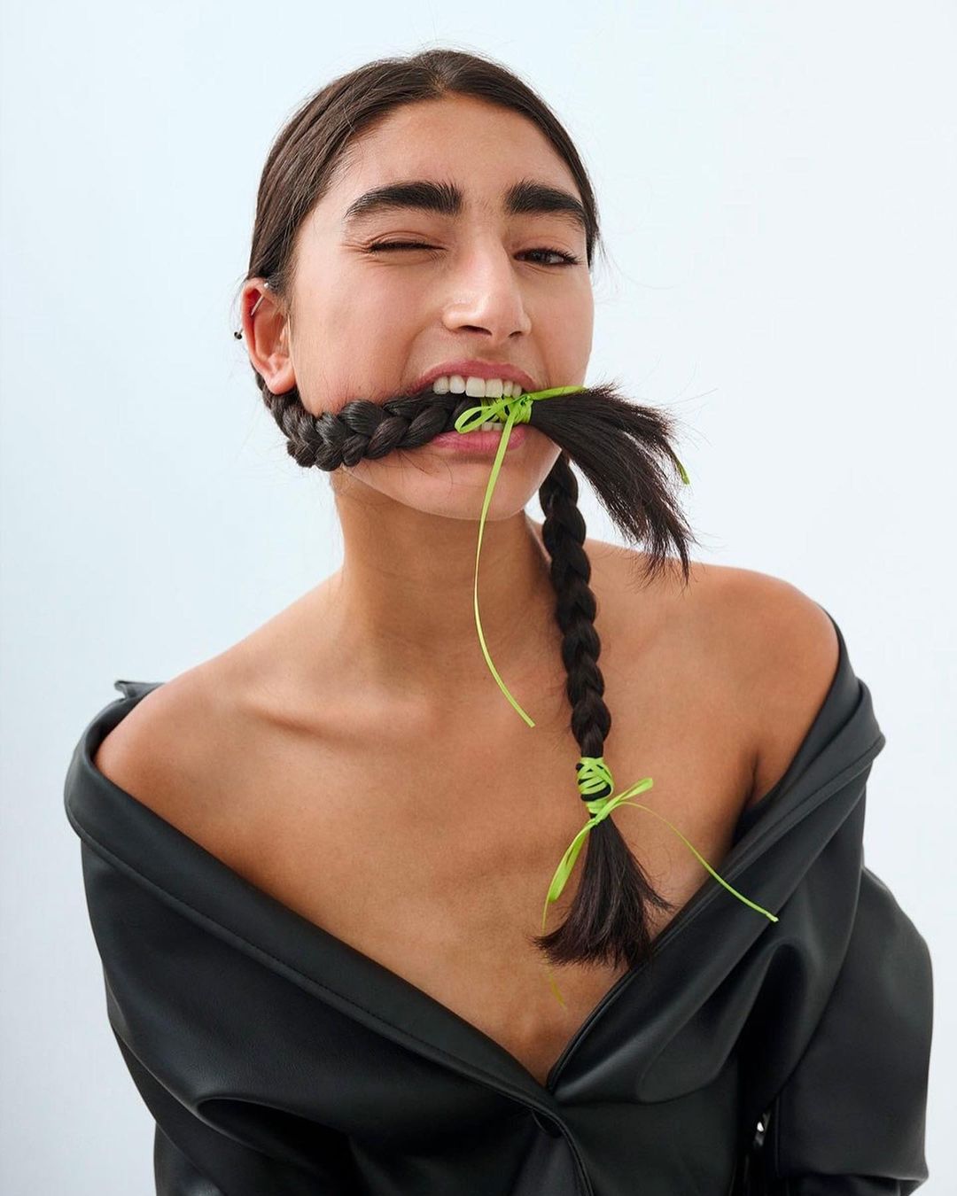 A young adult woman with dark hair in braids, wearing a black jacket against a white background, playfully winks at the camera.
