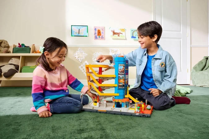 Two smiling children, a boy and a girl, are playing with a Matchbox toy garage on a green carpet inside a brightly lit room.