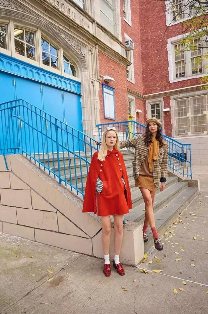 Two women in vintage-inspired outfits pose on school steps. One wears a red cape, the other a brown blazer and beret.