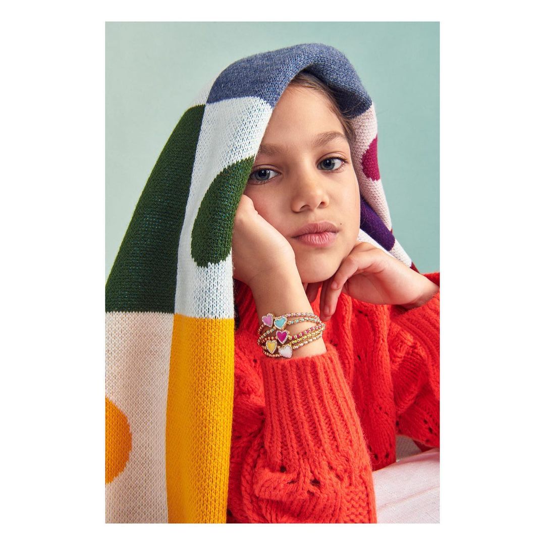 A young girl with fair skin and light eyes rests her head on her hands, wearing a red sweater and colorful knit hood indoors.