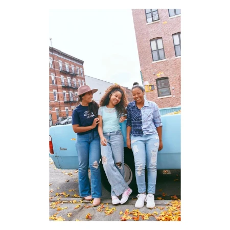 Three young women in denim fashion stand near a light blue truck with orange leaves on the ground against a brick building backdrop.