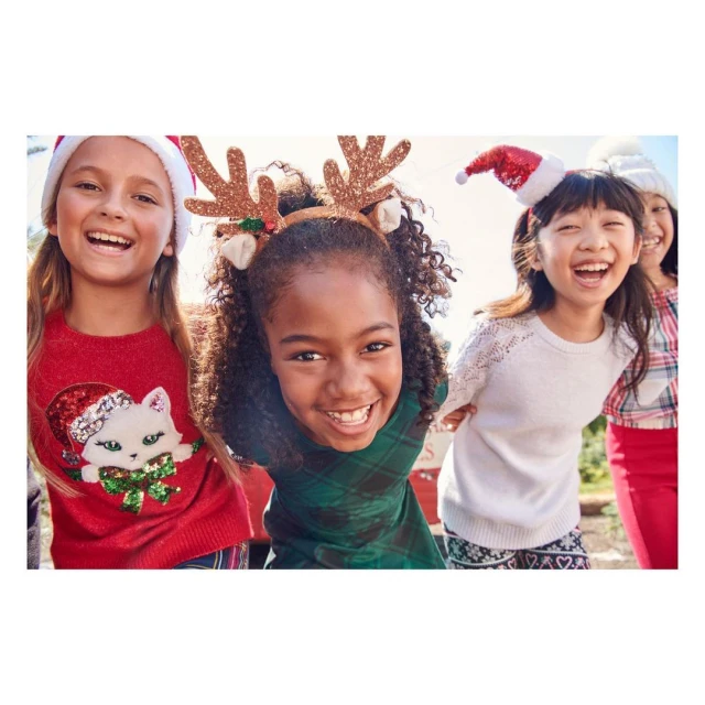 Four laughing girls wearing holiday headbands and sweaters pose outdoors. A joyful group portrait captures the festive spirit of the season.