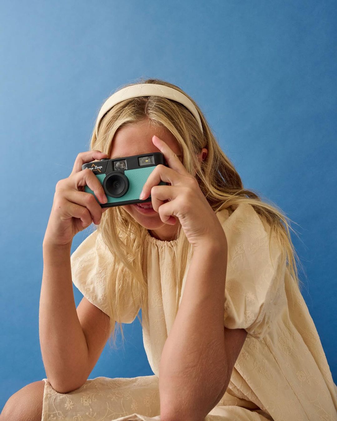A young female holds a vintage camera up to her face, smiling slightly, set against a solid blue background with soft, diffused lighting.