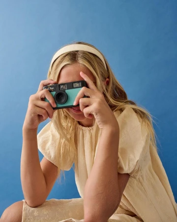 A young female holds a vintage camera up to her face, smiling slightly, set against a solid blue background with soft, diffused lighting.